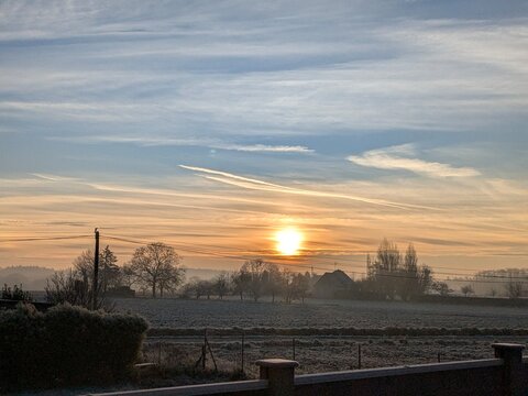 Lever de soleil hivernal au-dessus d&rsquo;un paysage de campagne recouvert de givre, sous un ciel nuageux aux teintes douces et chaleureuses, cr&eacute;ant une atmosph&egrave;re paisible presque picturale.