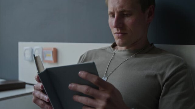 Zoom out rack focus shot of injured Caucasian young adult soldier with bandaged arm sitting in hospital bed and reading book. Clip for veteran rehabilitation and mental health recovery campaigns