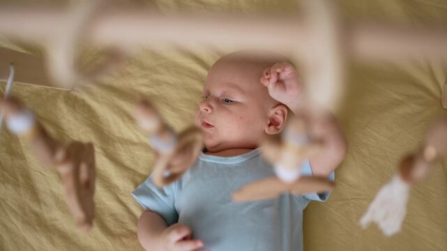 Adorable newborn baby lying awake in his crib and curiously watching the hanging wooden mobile, a sweet and authentic moment of early childhood development and sensory stimulation at home