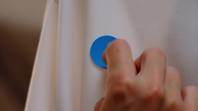 A hand carefully applies and presses a blue circular sticker onto a clean white garment, ensuring it adheres firmly before pulling away.