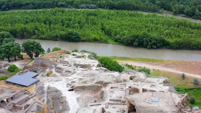 Drone flies forward above Uplistsikhe in Georgia, crossing pale rock terraces toward the Kura River and green woodland for archaeological tourism, conservation media and regional travel projects.