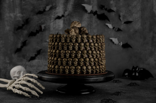 Halloween chocolate cake decorated with chocolate skulls and a skeleton hand on a black cake stand. Spooky ghost, pumpkin, and bat decorations create a dark festive atmosphere.