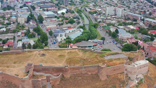 Aerial drone flies forward toward Gori Fortress in Georgia, approaching hilltop walls above busy streets and neighborhoods for landmark tourism, city branding and regional travel advertising.