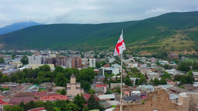 Georgian flag fills circling aerial view above Gori rooftops, church towers and surrounding hills, supporting city tourism, civic identity, cultural geography and Georgian destination campaigns.