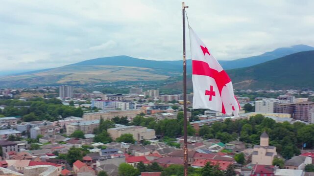 Drone circles beside the Georgian flag above Gori Fortress, revealing the fortified hill and city below for national heritage, cultural tourism, patriotic media and regional destination advertising.