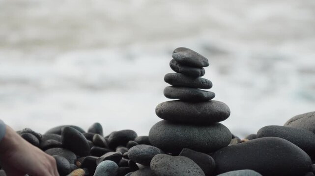 A person stacking pile of rocks on a beach, gentle wave in the background