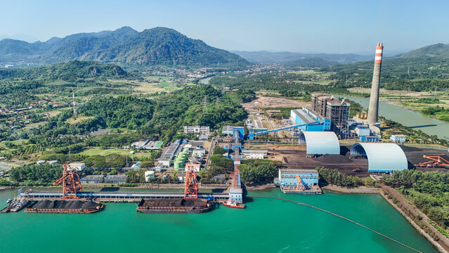 Aerial view of a coal-fired power plant with a tall chimney, coal storage domes, and a river terminal with barges in Indonesia.