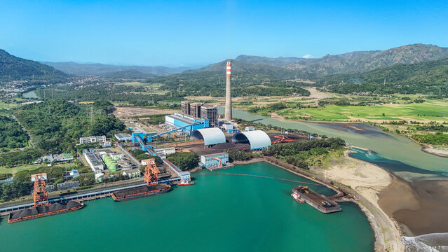 Aerial view of a coal-fired power plant with industrial chimneys, large storage domes, and coal barges docked at the coastline in Indonesia.
