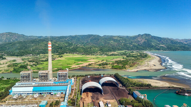 Aerial view of a power plant with a tall chimney and coal storage domes situated near the coast with green fields and mountains in the background, Indonesia.
