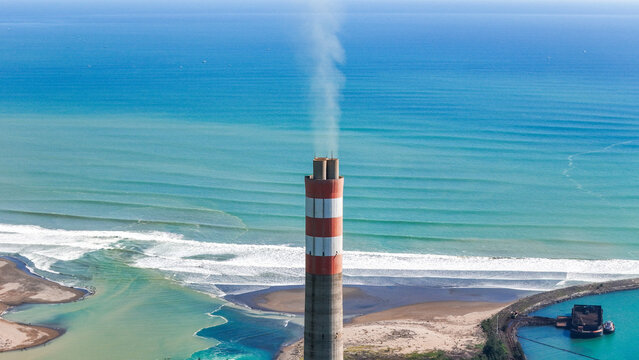 Aerial view of a tall red and white striped power plant chimney emitting smoke on the coastline with blue ocean waves and a small harbor in Indonesia.