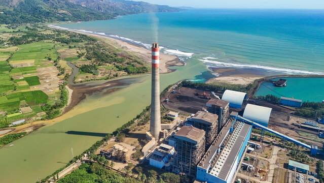 Aerial view of a power plant with a tall chimney situated near the coastline, surrounded by lush green rice fields and the blue sea in Indonesia.