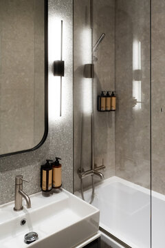 Modern bathroom interior featuring a clean washbasin, faucet, mirror, and a sleek vertical light, set against gray stone look tiles with a bathtub and shower in the background