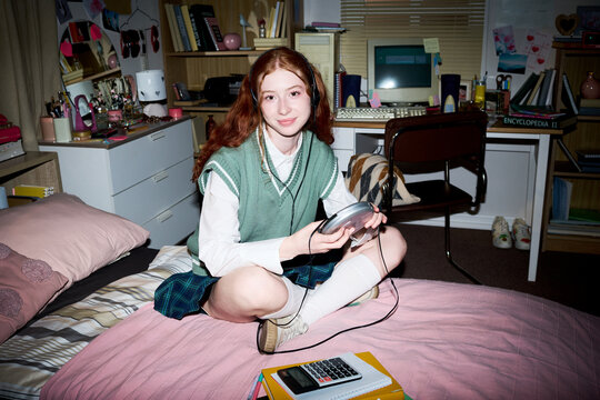 Portrait of Caucasian teenage girl sitting on bed, listening through headphones and holding portable CD player in bedroom. Useful for youth lifestyle, nostalgia, home media marketing