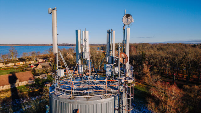 Valmiera, Latvia - 11 November 2025: Aerial view of a telecommunications tower equipped with various antennas and satellite dishes, set against a rural landscape with a lake and trees.