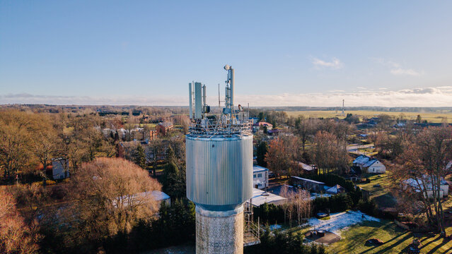 Valmiera, Latvia - 11 November 2025: Aerial view of a telecommunications tower with antennas and satellite dishes rising above a rural landscape with trees and buildings under a clear sky.