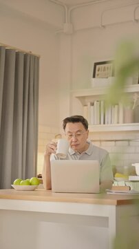 An elderly Asian man sits at the kitchen counter with a laptop and coffee mug, focusing on work or browsing during a calm morning at home. healthy retirement lifestyle, daily routine.