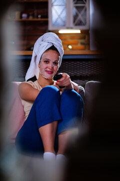 Young woman sitting comfortably on a sofa at home, wearing a white towel on her head and a beauty face mask, enjoying a moment of self care and relaxation while holding a remote control
