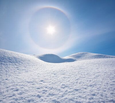 Beautiful winter landscape with a spectacular sun halo effect over smooth white snow hills under a blue sky.
