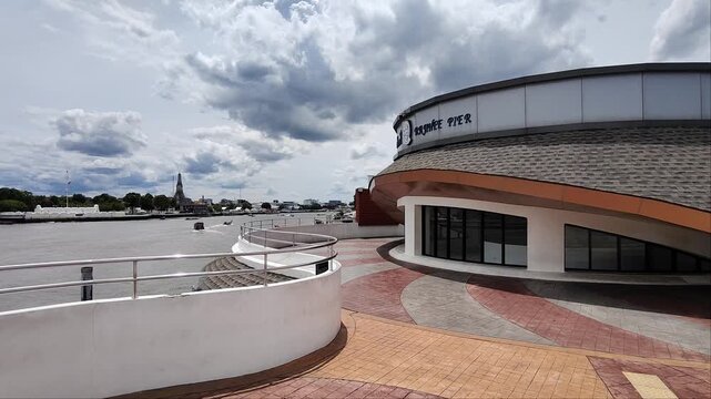 Scenic view of the curved exterior of Rajini Pier and riverside promenade in Bangkok, Thailand, with dramatic clouds and river traffic in the background.