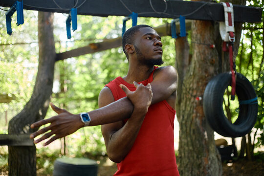 Medium shot of young Black man stretching arm before outdoor obstacle training, preparing body for fitness challenge and active lifestyle marketing or wellness campaign