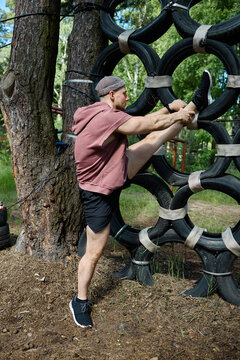 Vertical shot of Caucasian young adult man climbing tire obstacle during outdoor fitness training, stretching legs and hamstrings. Ideal for boot camp promotion, wellness marketing