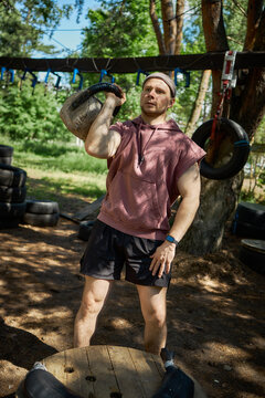 Vertical shot of Caucasian young adult man lifting kettlebell on shoulder during outdoor functional workout, building strength and endurance. Fitness marketing, personal training, bootcamp promotion