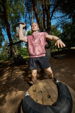 Vertical shot of young adult Caucasian man training with kettlebell outdoors, building strength and endurance in obstacle course setting. Fitness marketing, bootcamp promotion, wide angle shot