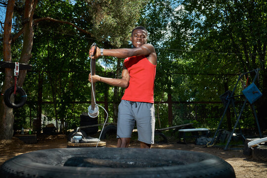 Young Black man training outdoors, swinging weighted rope beside tire and building strength for functional fitness. Useful for sports marketing, workout programs, resilience themes