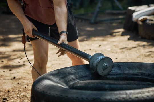 Young adult man gripping sledgehammer and striking tire during outdoor workout, showing strength training and functional fitness. Useful for gym promotion, bootcamp ads, wellness content