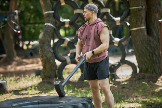 Caucasian young adult man training with sledgehammer on tire in outdoor obstacle gym, building strength and endurance. Medium full shot, fitting fitness bootcamp or workout promotion