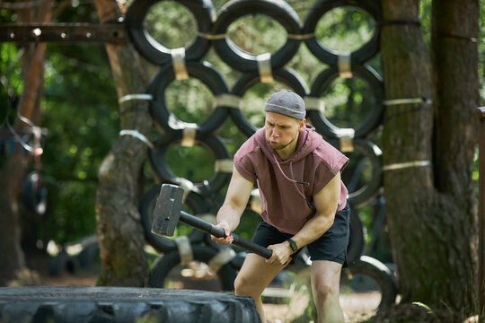 Young adult Caucasian man training with sledgehammer on tire at outdoor obstacle course, building strength and endurance. Wide shot, fitting fitness bootcamp or resilience marketing