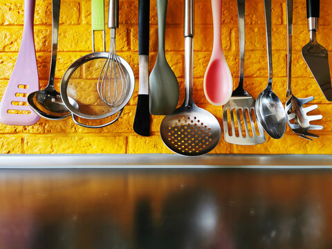 Bright kitchen utensils hanging on a railing against a yellow brick wall&mdash;a spatula, a slotted spoon, tweezers, a ladle, a whisk, a sieve, and a spoon. Silicone and metal utensils