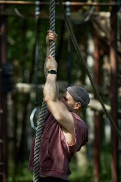 Vertical shot of young adult Caucasian man climbing rope during outdoor obstacle training, building strength and endurance. Useful for fitness marketing, bootcamp promotion, wellness content