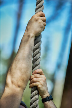 Vertical shot of Caucasian young adult man gripping climbing rope while ascending outdoor obstacle course, conveying strength and determination. Ideal for fitness, teamwork, challenge marketing