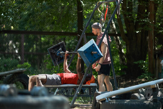 Black young adult man lifting improvised barbell on outdoor bench while Caucasian young adult trainer supervising workout, promoting fitness coaching, strength training marketing