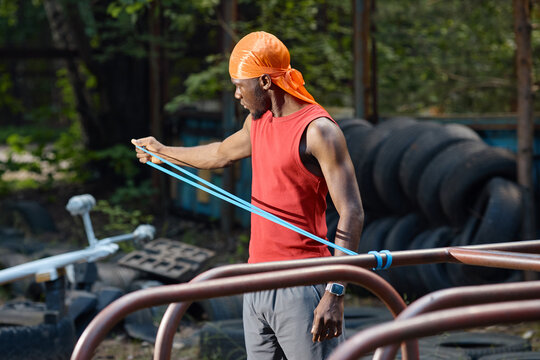 Black young adult man stretching resistance band during outdoor workout, building strength and mobility in urban fitness space. Ideal for sports training, wellness, active lifestyle marketing