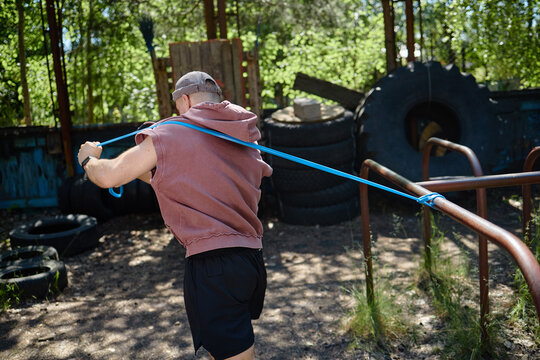 Young adult man training with resistance band in outdoor fitness area, strengthening upper body and endurance. Useful for sports coaching, workout programs, healthy lifestyle marketing
