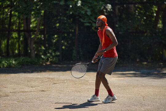 Full body shot of young adult Black man playing tennis on outdoor court, preparing return swing and training for active lifestyle concept. Ideal for sports marketing, fitness campaigns
