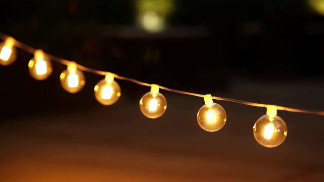 Diagonal string lights glowing at dusk with warm filament reflections in clear globe bulbs suspended on dark wire for cozy outdoor decor, with bulb, glass, string, wire detail