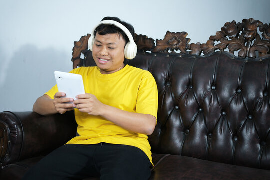 A relaxing weekend at home. A happy and smiling young Asian man spends his free time relaxing on the living room couch, playing on his phone, listening to music with his headphones, reading blogs on s