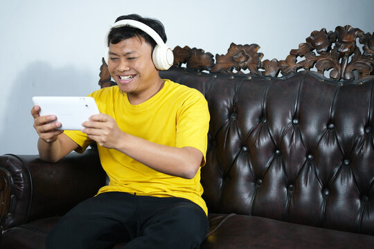 An Asian man focused on playing a game on his smartphone, while lying on the sofa with a big smile on his face, having won the game. Enjoying the weekend by relaxing and playing games.
