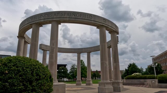 Low Angle View of University Stone Colonnade Arch on Campus Quad. Ground-level shot of stone arch on college quad with modern buildings and dynamic clouds behind. At University of Illinois Springfield