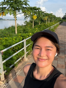 Happy Thai woman in sportswear taking a selfie on a riverside path with green trees.