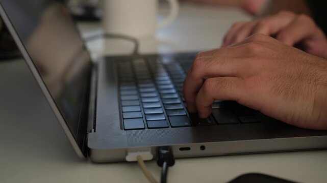 Close Up of Man Hands Typing on Laptop Keyboard. Side view close up of male hands typing on a laptop keyboard at a white table while working online in a dimly lit indoor space.