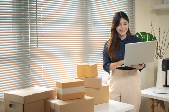 Young Asian Woman Entrepreneur Managing Online Orders with Laptop Surrounded by Cardboard Boxes at Home Office