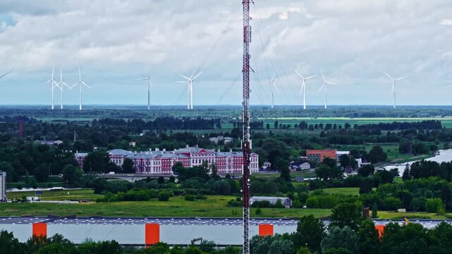 Aerial view of Jelgava Palace surrounded by lush greenery, wind turbines, and a tall radio tower under a cloudy sky in Jelgava, Zemgale, Latvia.