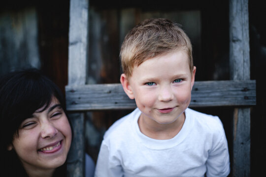 Portrait of two cheerful siblings standing together in front of a rustic wooden house with playful smiles and natural expressions