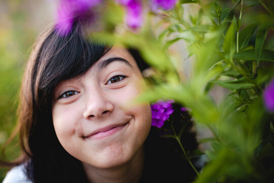 Close up portrait of a smiling young girl peeking through purple flowers and green leaves in a blooming summer garden
