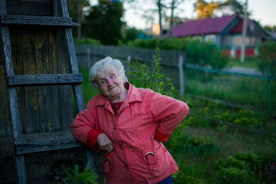 Environmental portrait of a smiling elderly woman leaning beside a weathered wooden ladder in a quiet rural garden at sunset