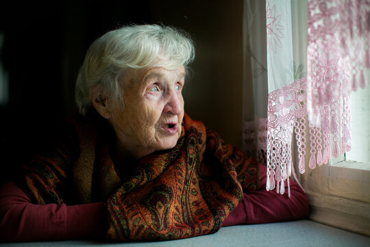 Portrait of an elderly woman wrapped in a warm patterned shawl looking thoughtfully through a lace curtained window in soft natural light